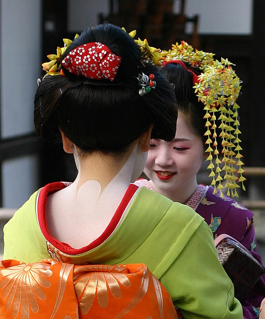 A photograph of 2 Geisha conversing near the Golden Temple in Kyoto, Japan. Parts of the kimono and the special make-up are clearly visible.